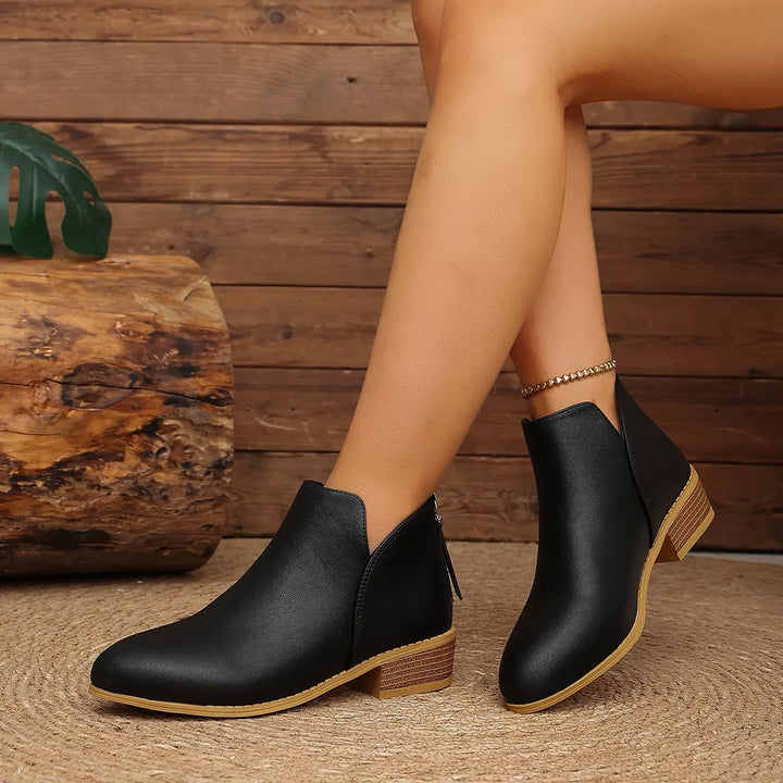 Women's ankle boots in black, low block heel with V-cut front, worn with legs crossed