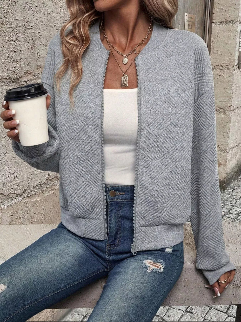 Light gray cropped bomber jacket styled with jeans, seated holding a coffee