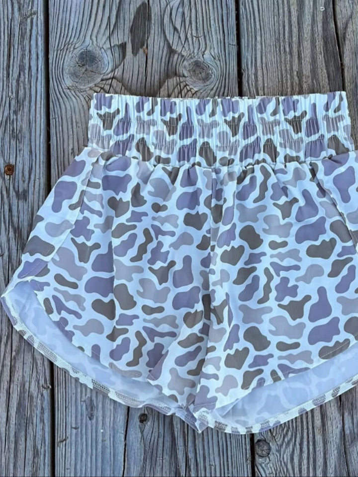 Women's camo shorts in blue camo, high waisted with tulip hem, laid flat on wood