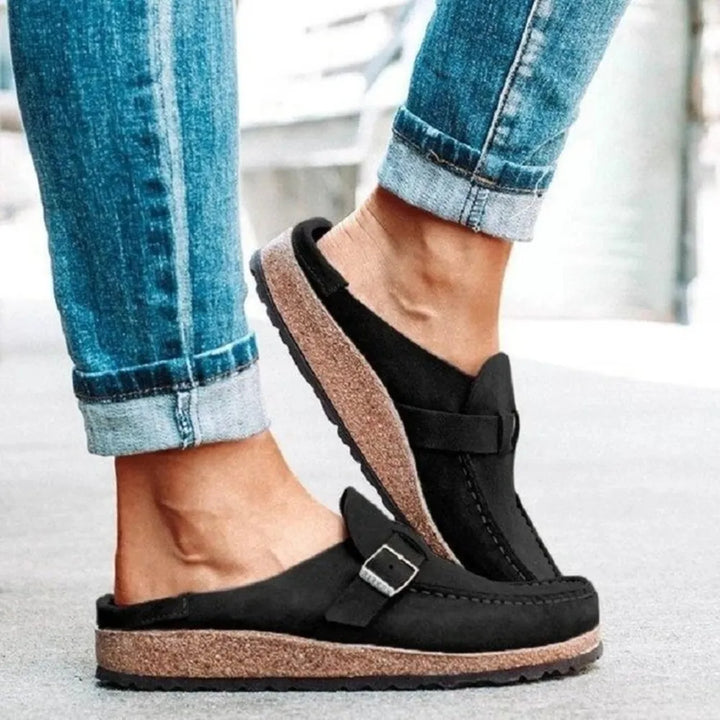 Black backless wedge clogs with buckle strap, side view while stepping
