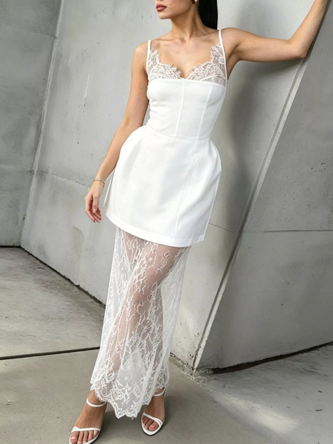 White corset mini dress with long lace overlay, leaning against a concrete wall