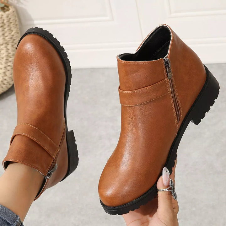 Cognac ankle boots held in hand showing the side zipper and buckle strap
