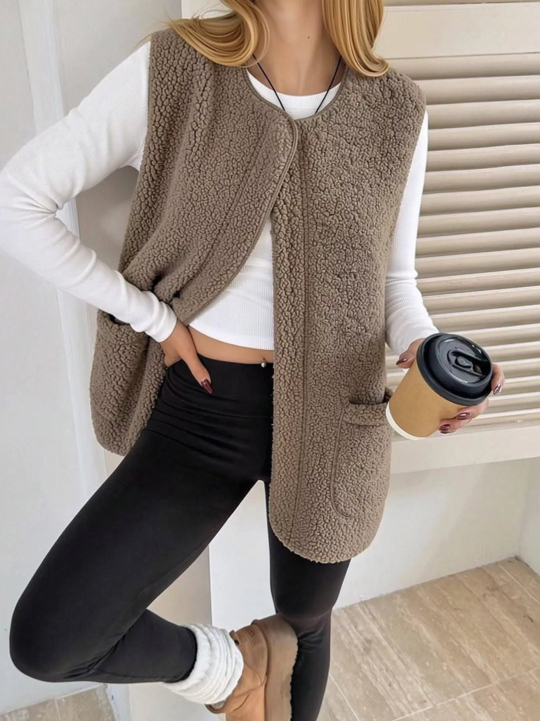 Taupe fuzzy vest worn open over a white top and black leggings with a coffee cup