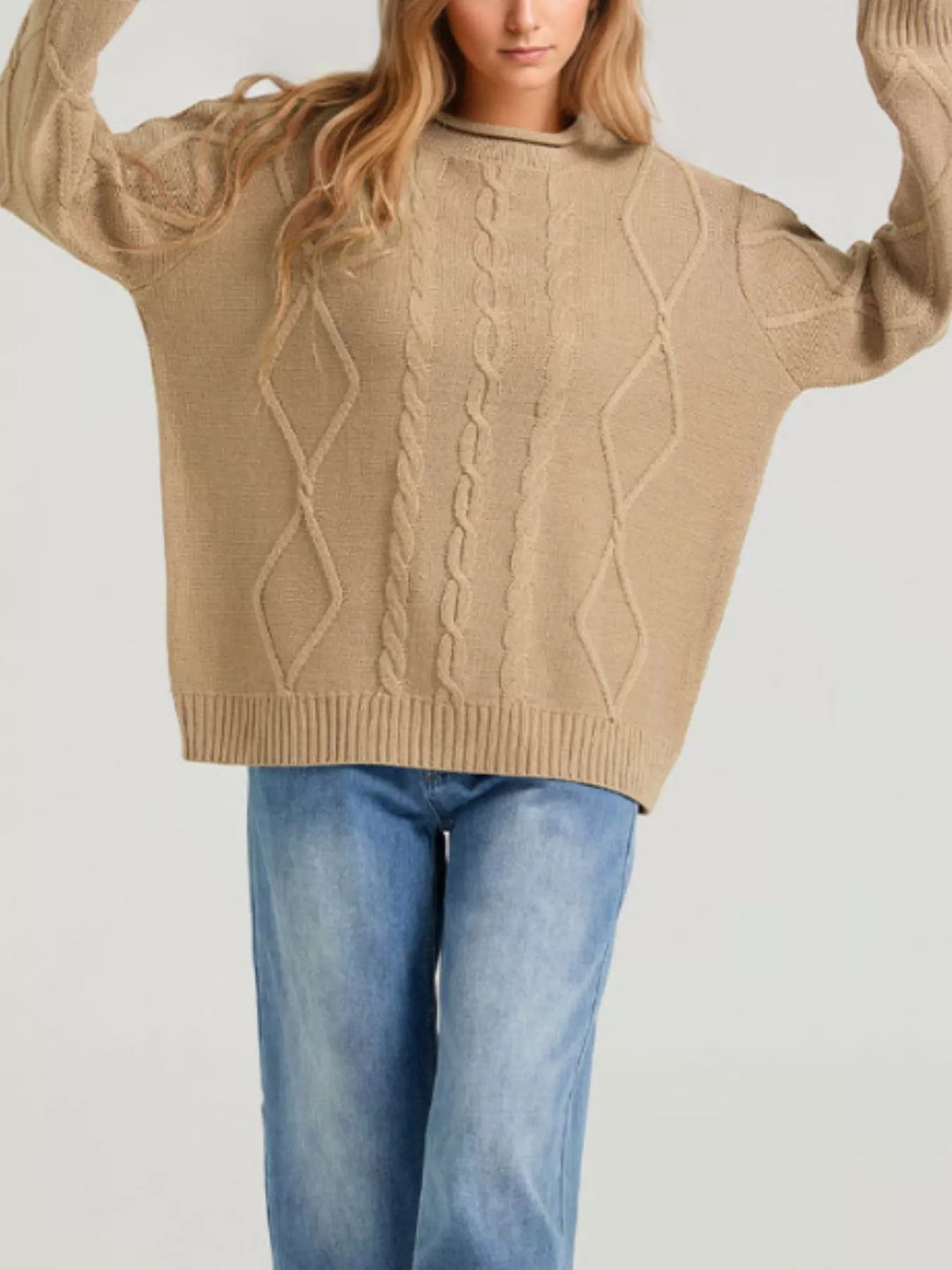 Tan cable knit sweater worn with arms raised over blue jeans