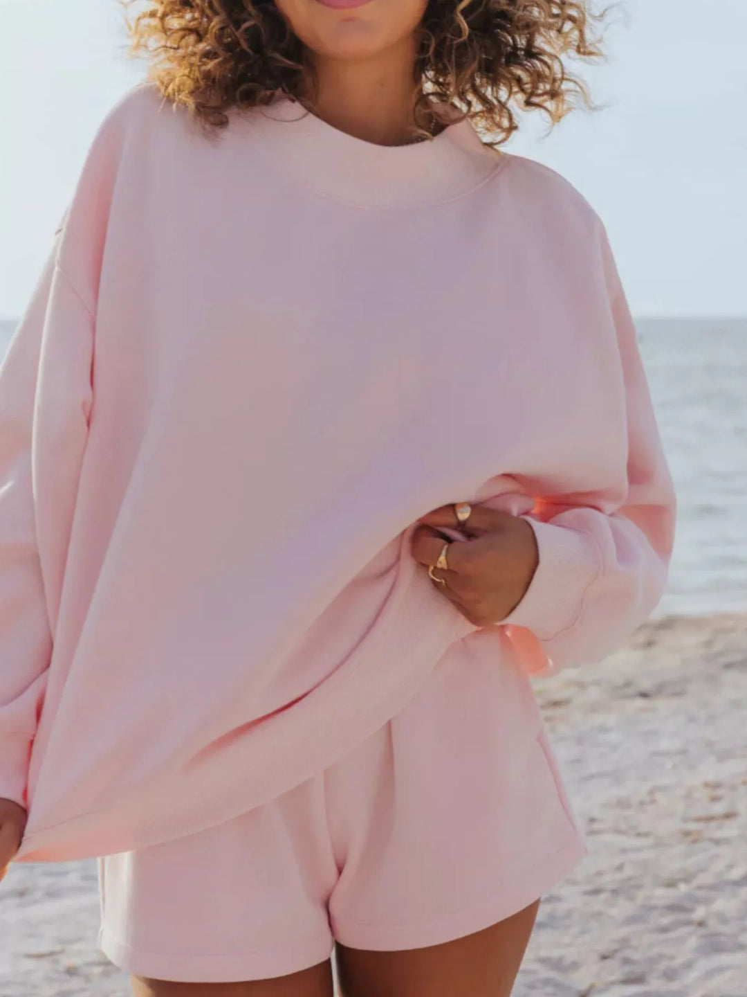 Blush pink oversized sweatshirt and shorts, relaxed side pose by the ocean