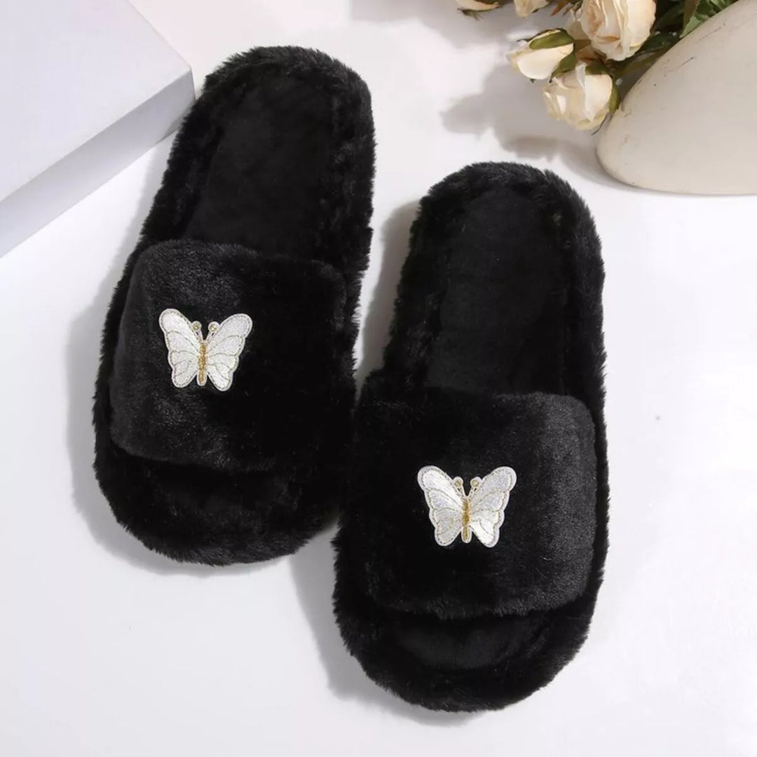 Pair of black fluffy slide slippers with butterfly embellishment on a white surface