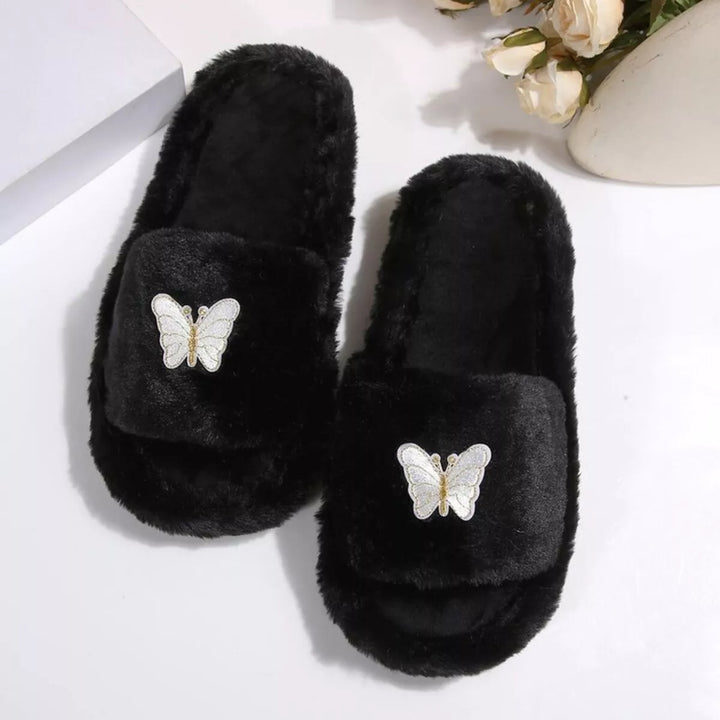 Pair of black fluffy slide slippers with butterfly embellishment on a white surface