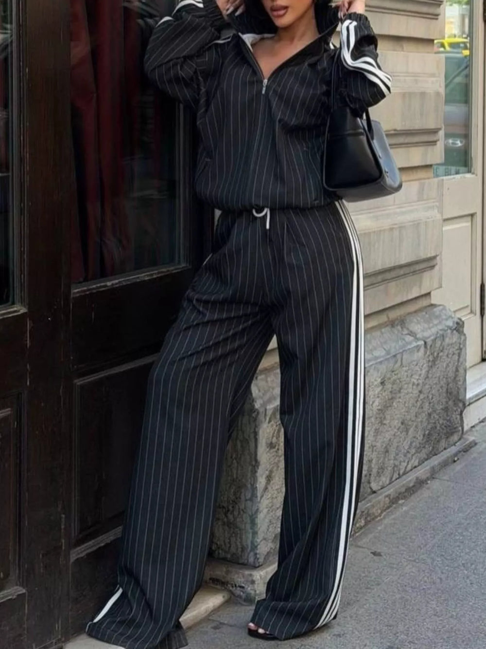 Black pinstripe zip jacket and wide leg pants, leaning on a doorway with a handbag