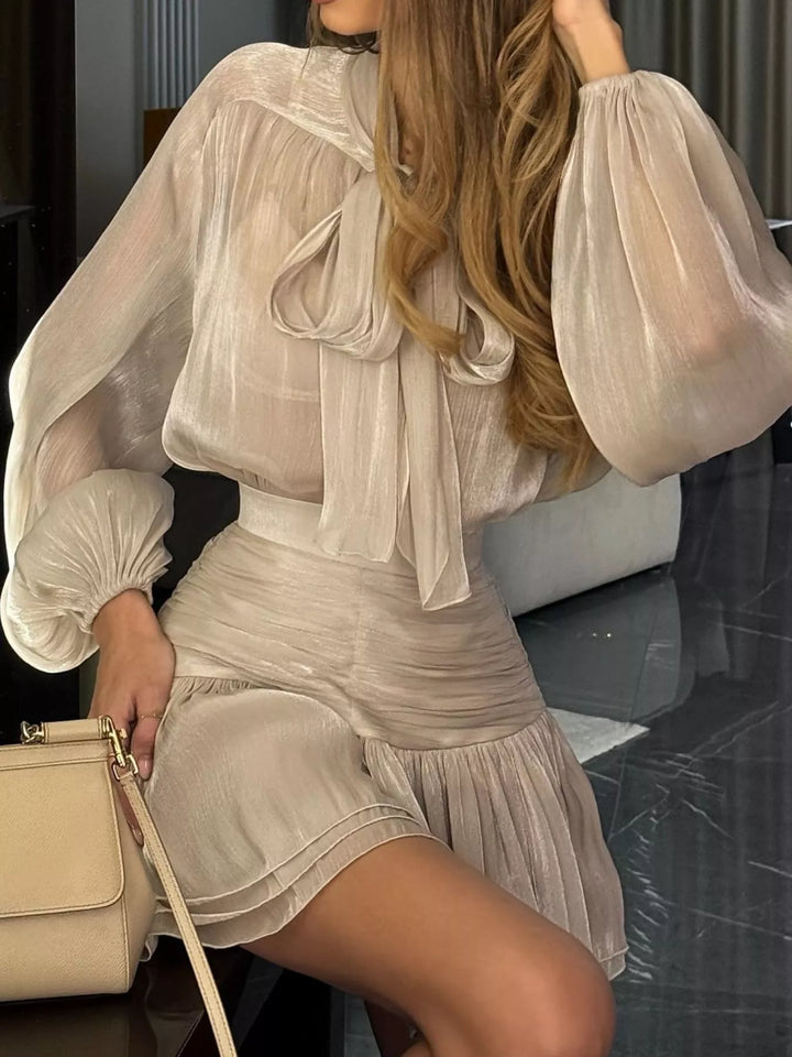 Champagne bow tie blouse and ruffle mini skirt set, seated with a beige handbag