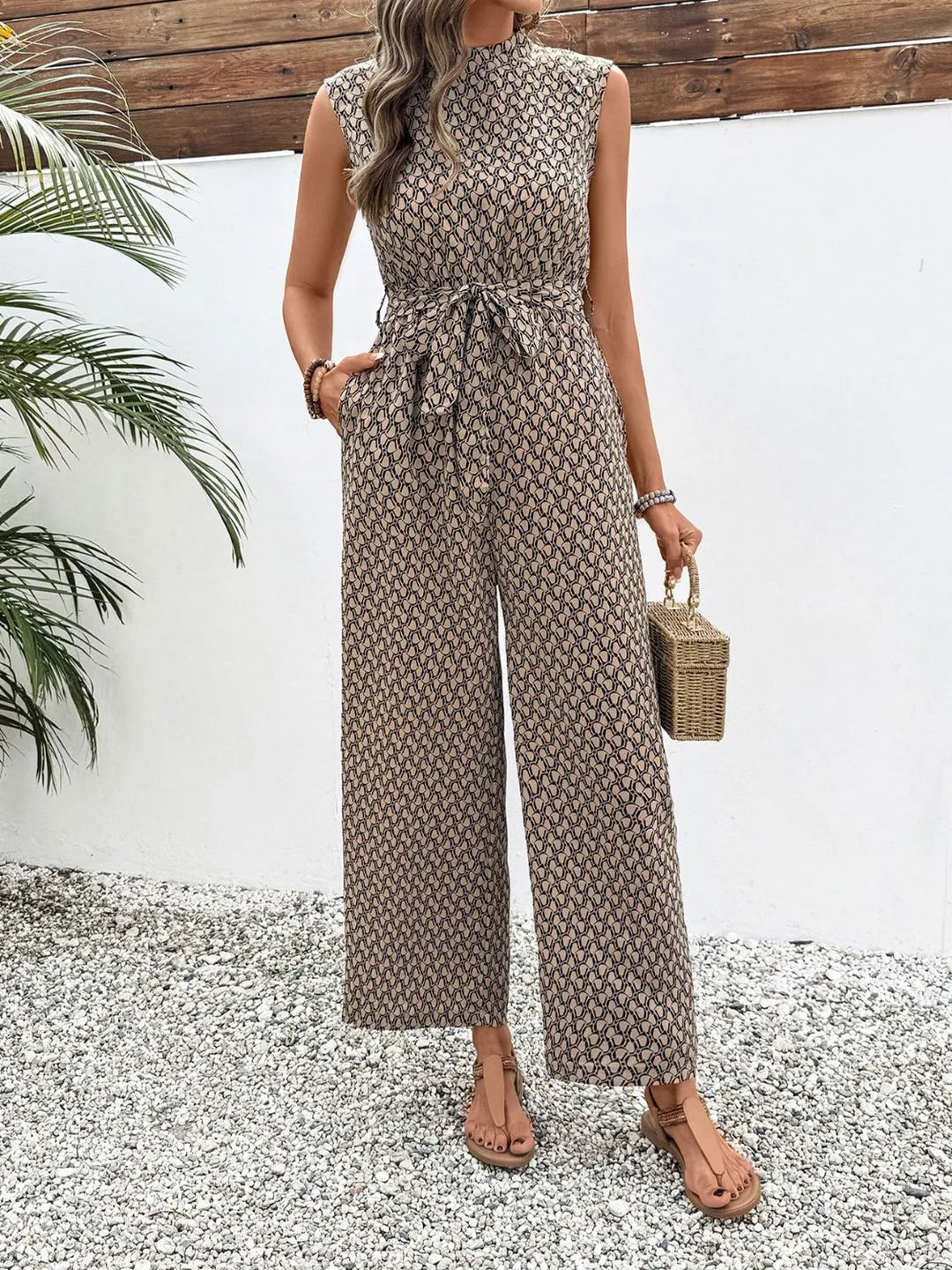 Full-length view of the taupe print jumpsuit with a woven straw bag in hand