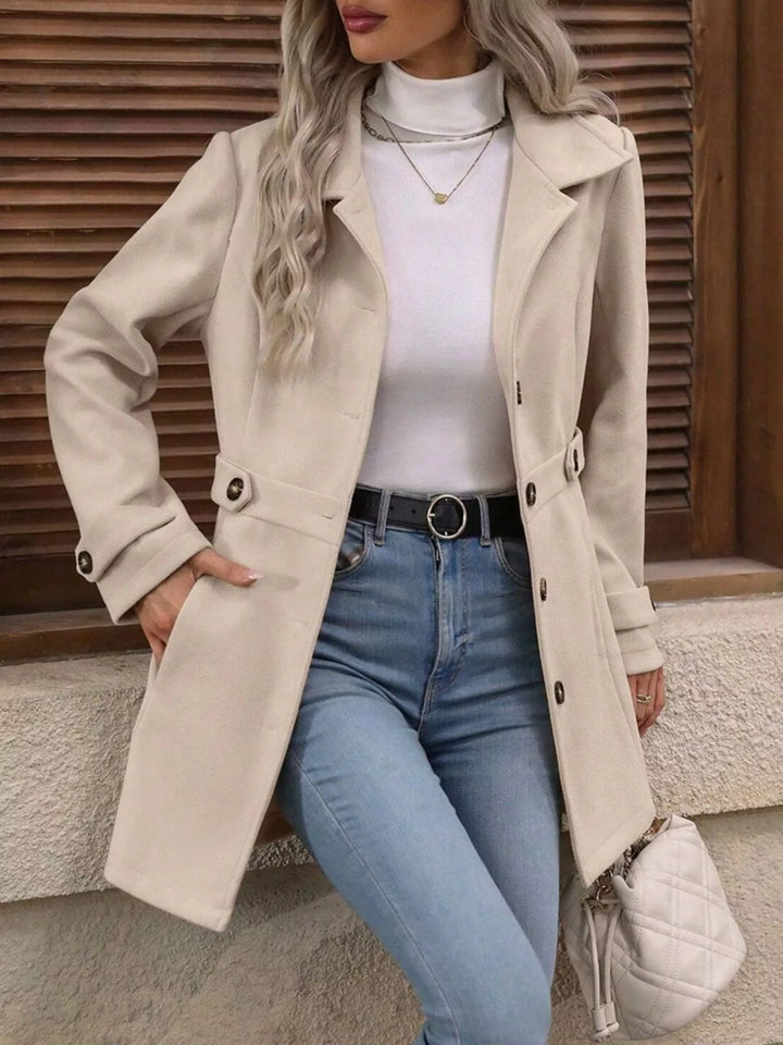 Beige collared coat worn open with hand on pocket, jeans and quilted bag
